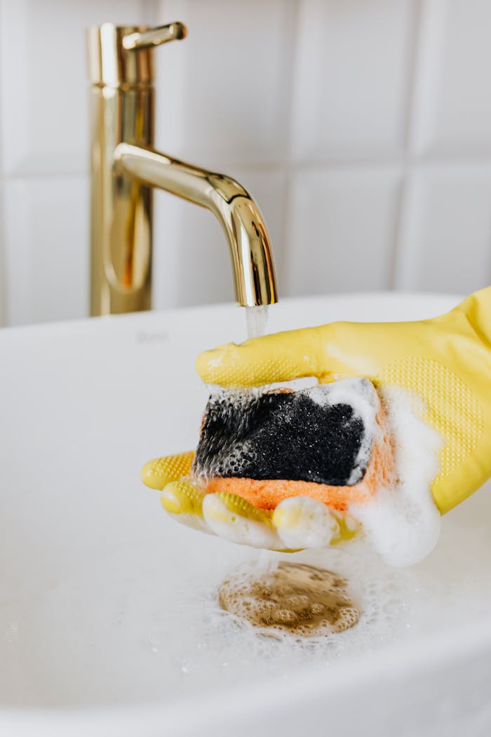 Close-up of a yellow-gloved hand cleaning with a foamy sponge under a gold faucet in a sink.